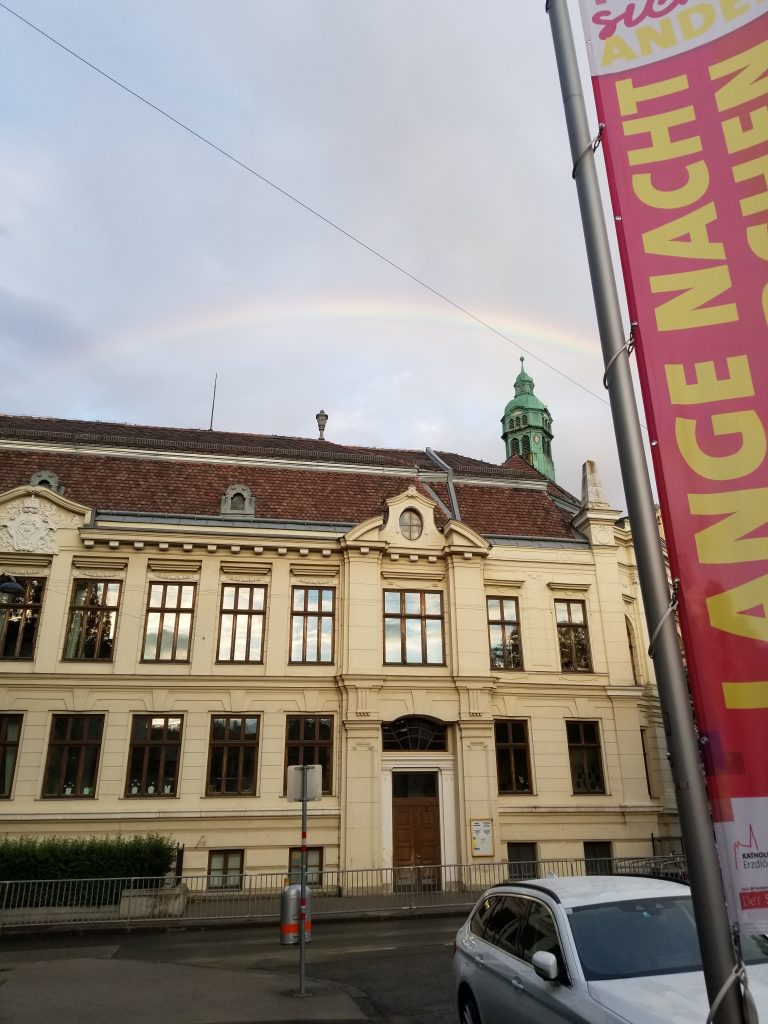 Ein Regenbogen ist am Himmel zu sehen über einem Gründerzeitschulhaus, eine Fahne am Bildrand auf der steht "Lange Nacht"